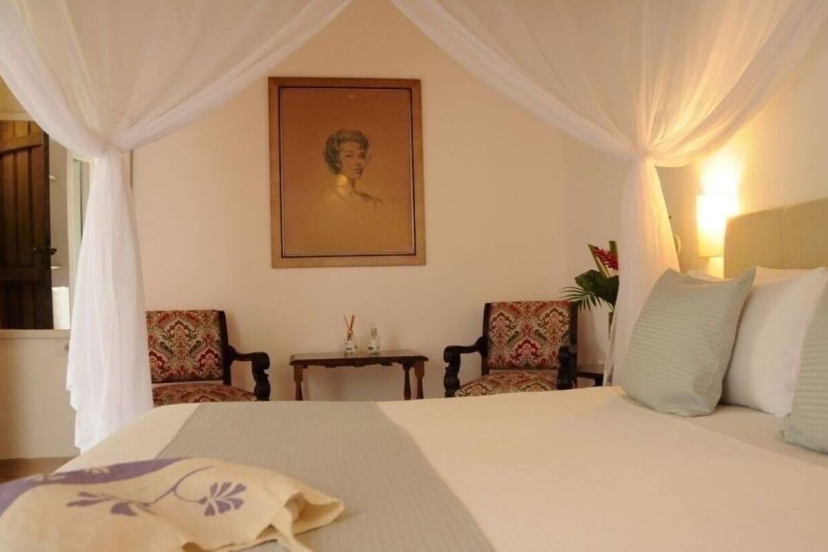 Un dormitorio con cama a dosel, dos sillones, una mesita y un retrato en la pared.