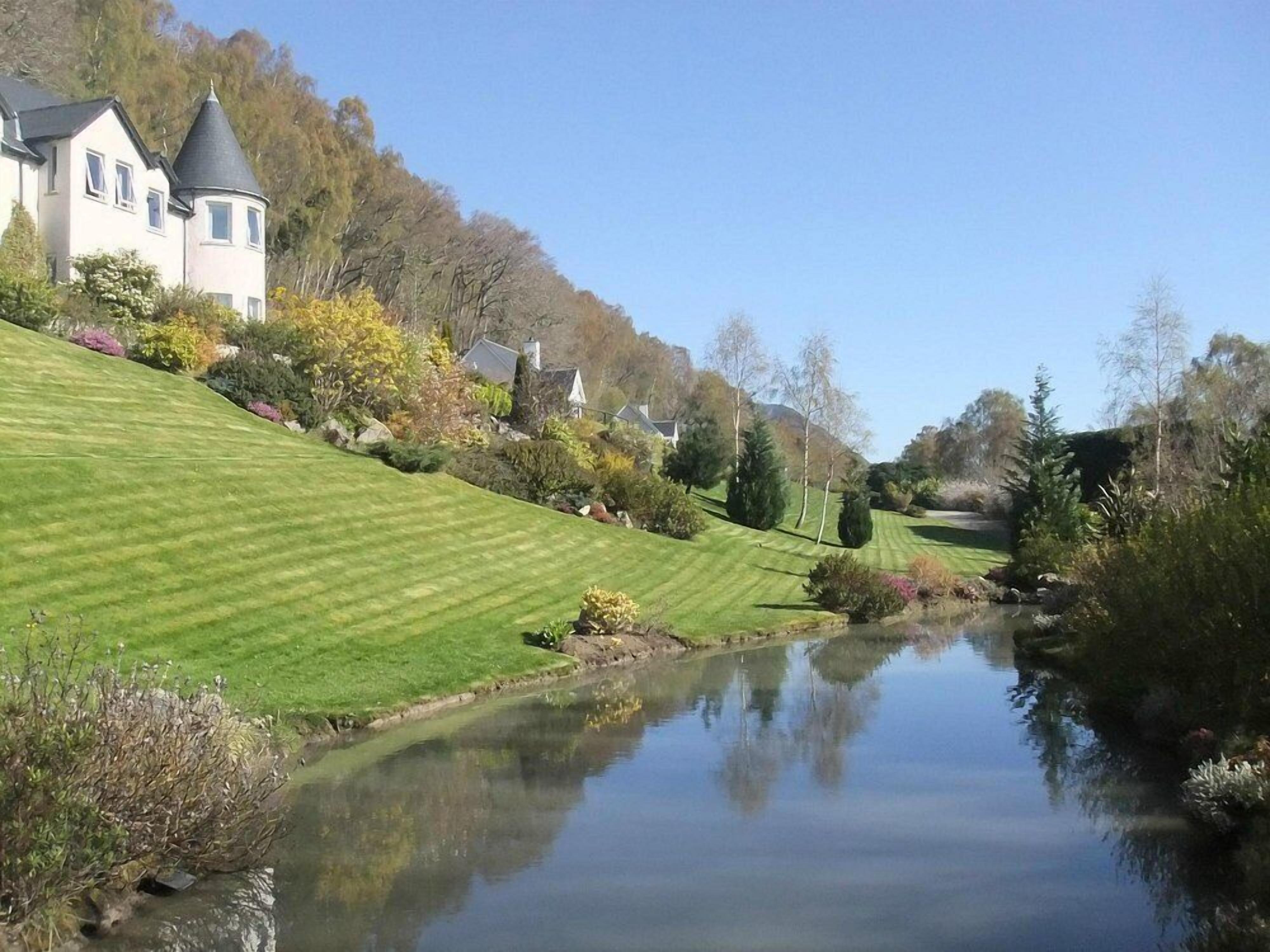 A well-maintained lawn with a pond reflecting the surroundings, a house with a turret, and a clear sky.