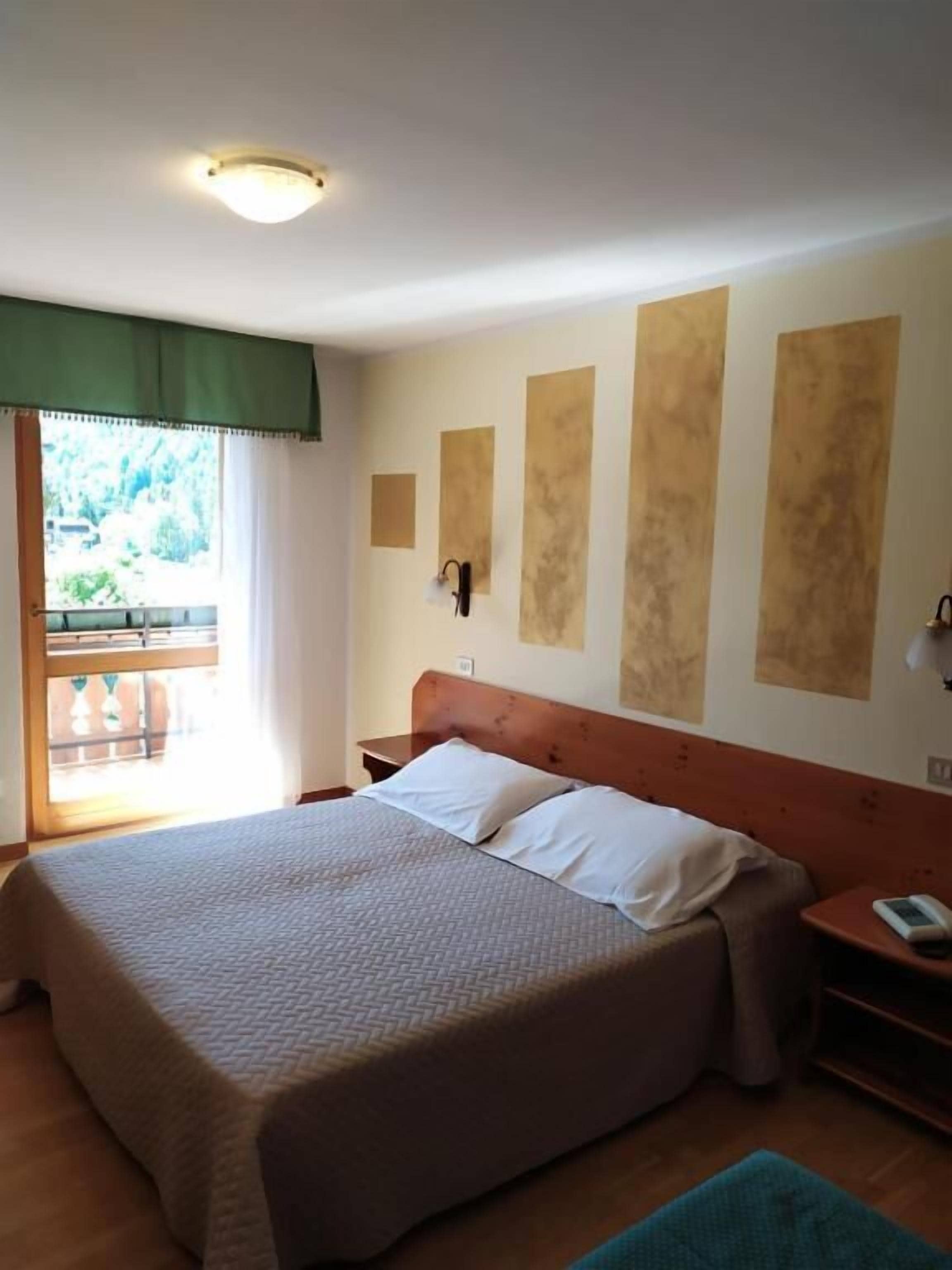 A hotel room with a large bed, a nightstand, a wall-mounted lamp, and a window with a view of greenery.