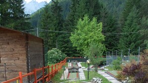 A wooden cabin with a red fence, a stone pathway, and a green mat leading to a seating area with plants and a tree. - Albergo Ristorante Cacciatori  (Auronzo di Cadore)