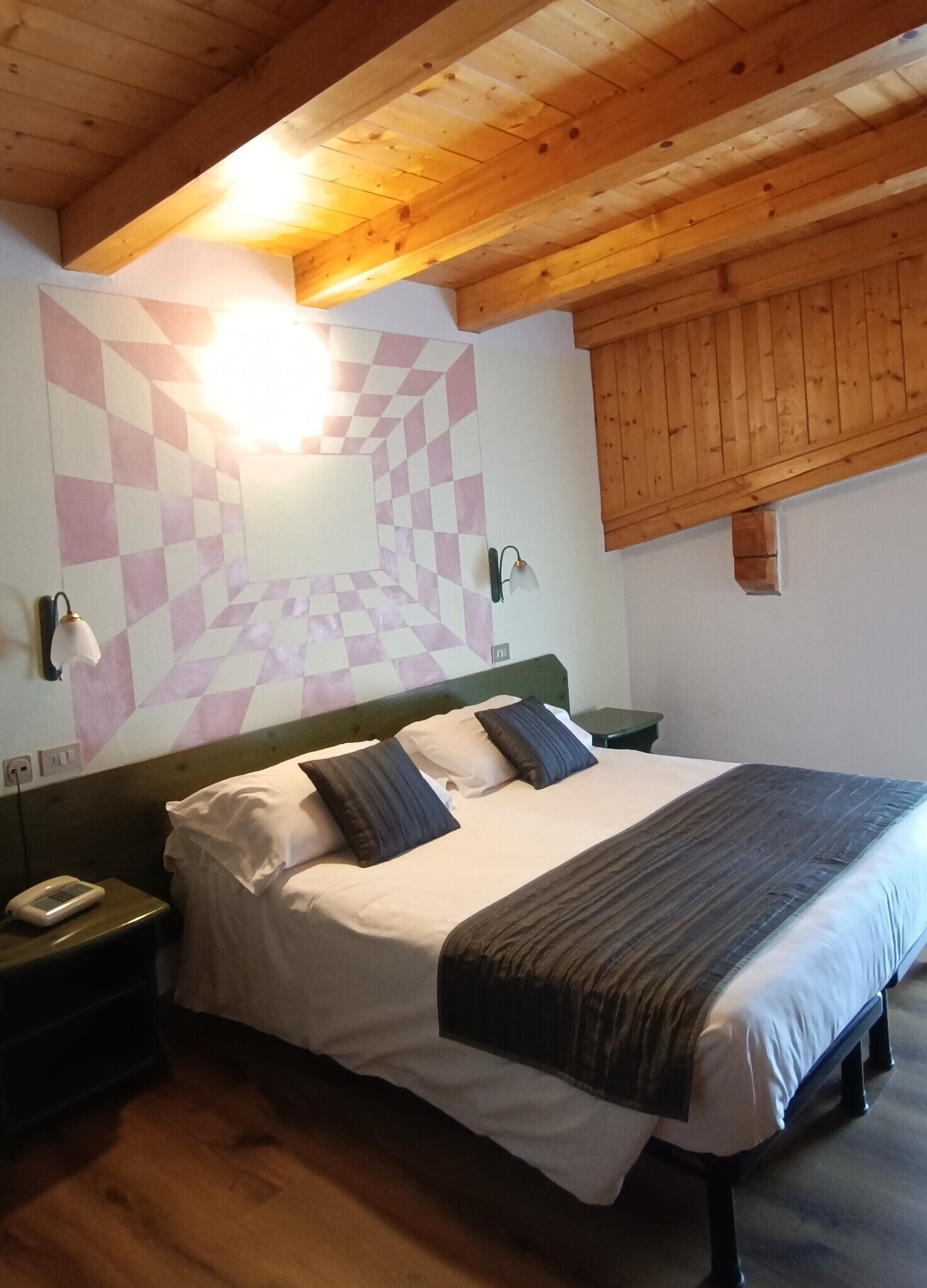 Un dormitorio con cama, cabecero de madera, mesitas de noche, una lámpara de pared y un diseño de pared a cuadros.