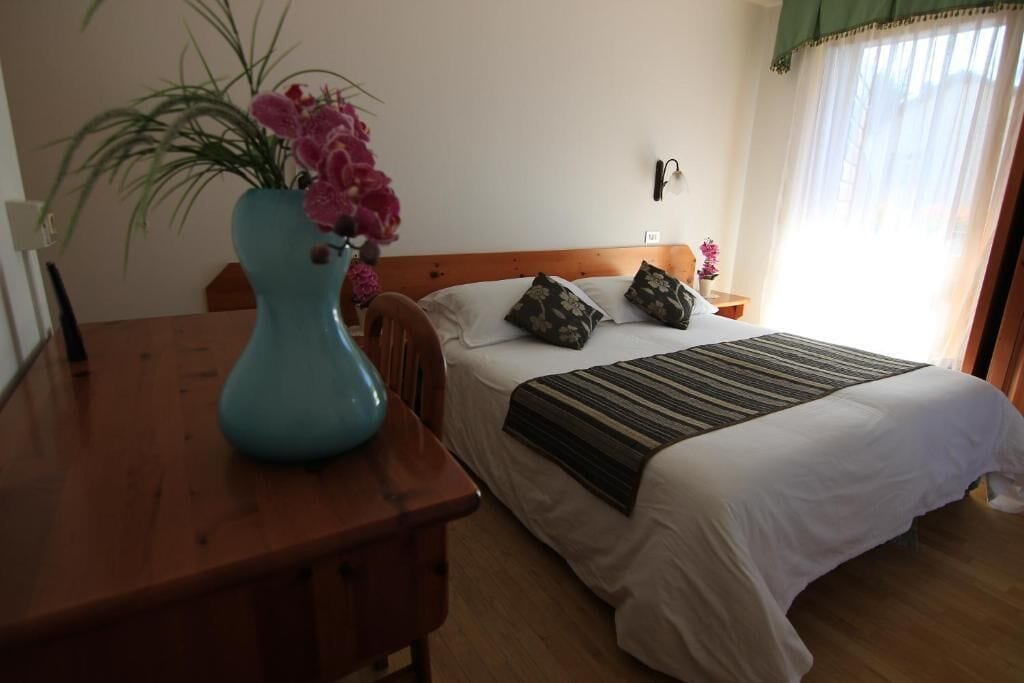 A bedroom with a wooden headboard, a bed with white linens, a striped blanket, and a vase with pink flowers on a wooden table.