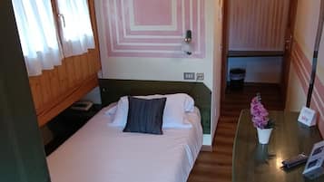 Ein Zimmer mit einem Bett, einem Schreibtisch und einem Stuhl. Es gibt ein Fenster mit Vorhängen, eine Wand mit geometrischem Muster und eine Holzbalkendecke.