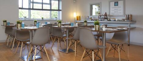 A modern dining area with multiple tables and chairs, large windows, and a counter with amenities.