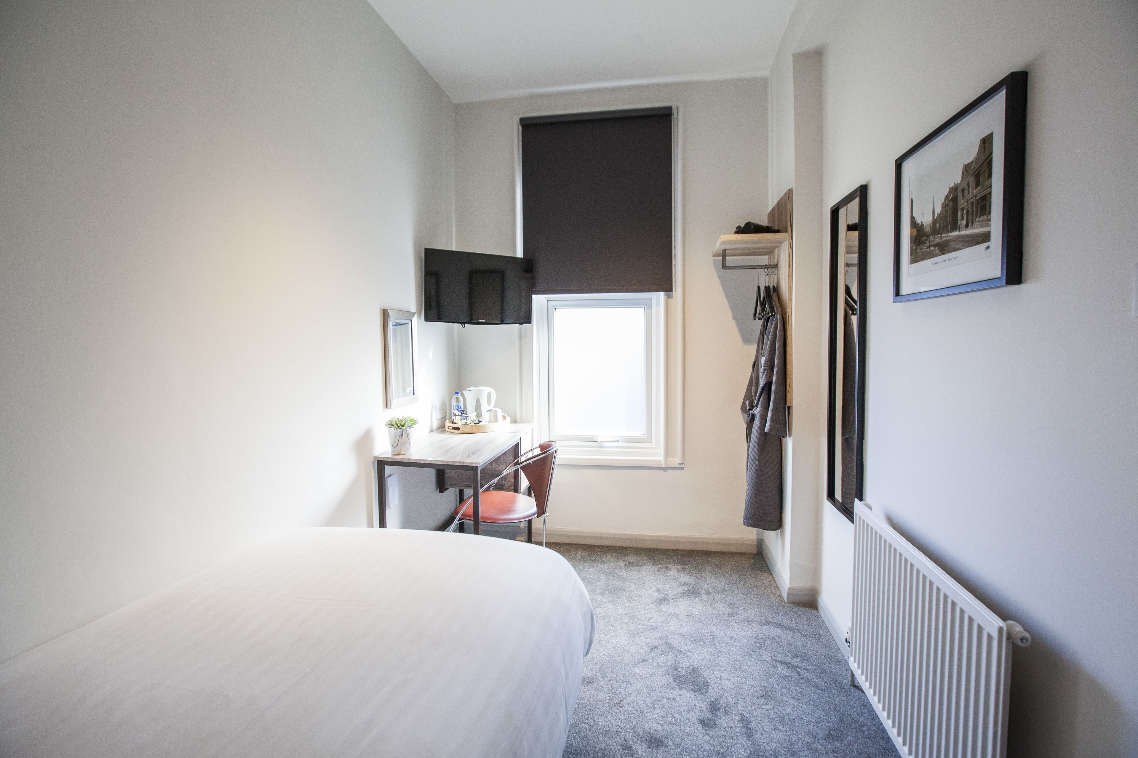 Una habitación de hotel con una cama, un escritorio con una silla, una ventana con persianas, un radiador y un cuadro en la pared.