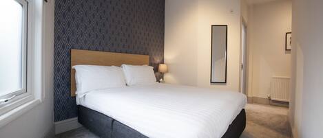 A bedroom with a large bed, a nightstand with a lamp, a window, and a wall with a geometric pattern.
