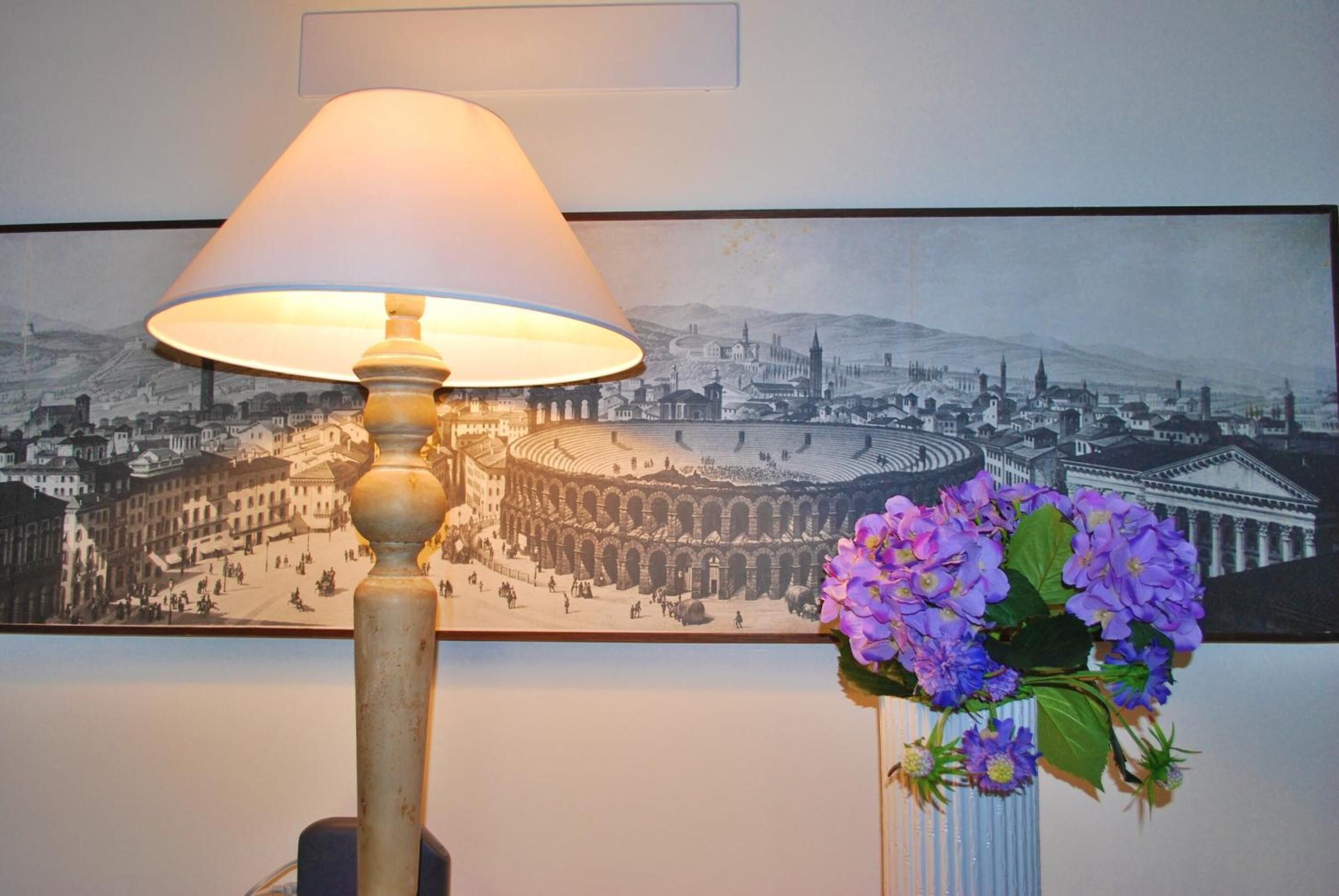 A lamp with a beige shade and a vase with purple flowers on a wall.