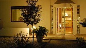 A modern building with a glass door entrance, a potted plant in the foreground, and a small garden area. - Villa Bianca (Sona)