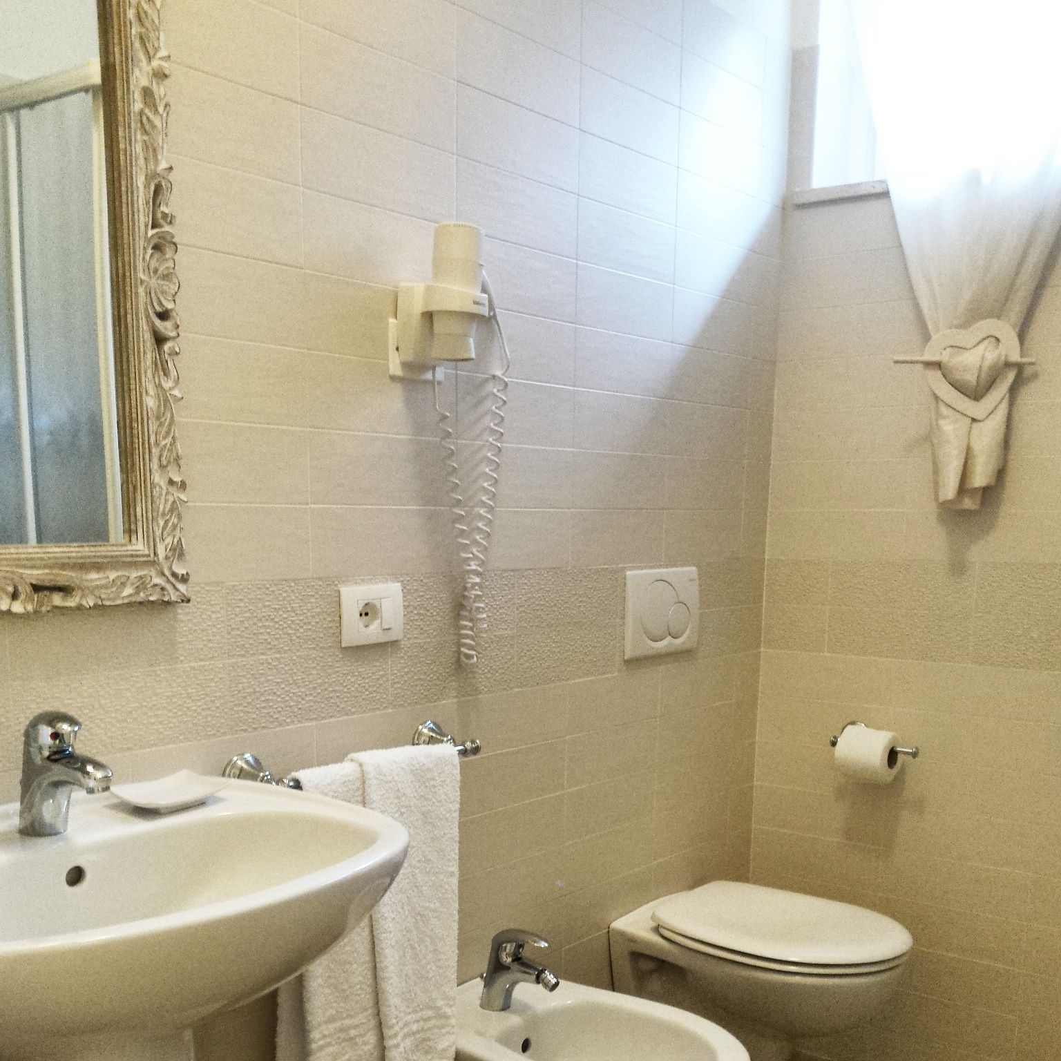 Un baño con lavamanos, inodoro y ducha. Se observa un espejo con marco decorativo, una toalla colgada en la pared y un perchero.