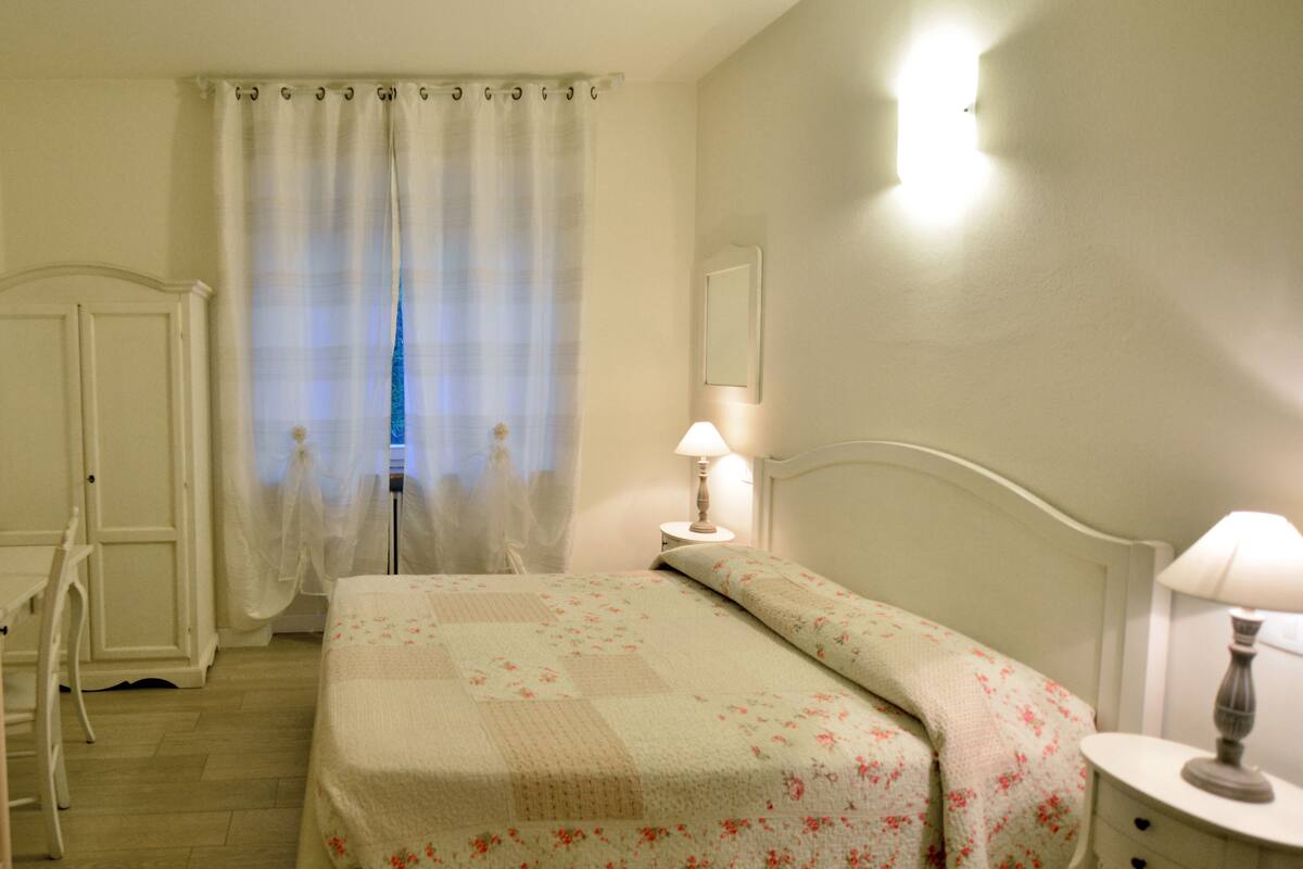 Ein Schlafzimmer mit einem Bett, zwei Nachttischen mit Lampen, einem Sessel, einem Kleiderschrank und einem Fenster mit Vorhängen.
