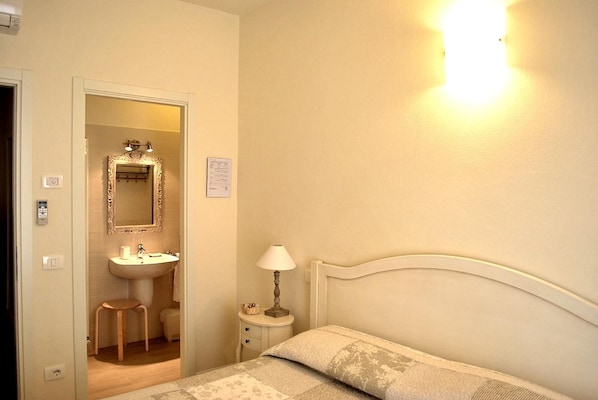 A bedroom with a bed, a bedside table with a lamp, a small stool, and a bathroom with a sink and mirror. - Villa Bianca (Sona)