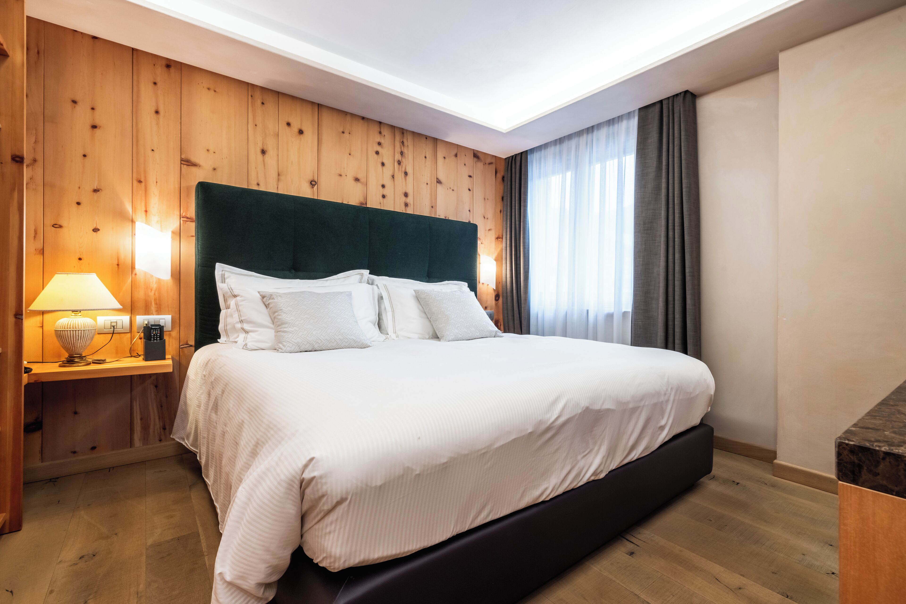 A hotel room with a wooden paneled wall, a large bed with white bedding, a bedside table with a lamp, and a window with curtains.