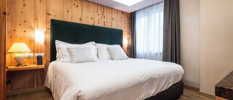 A hotel room with a wooden paneled wall, a large bed with white bedding, a bedside table with a lamp, and a window with curtains.