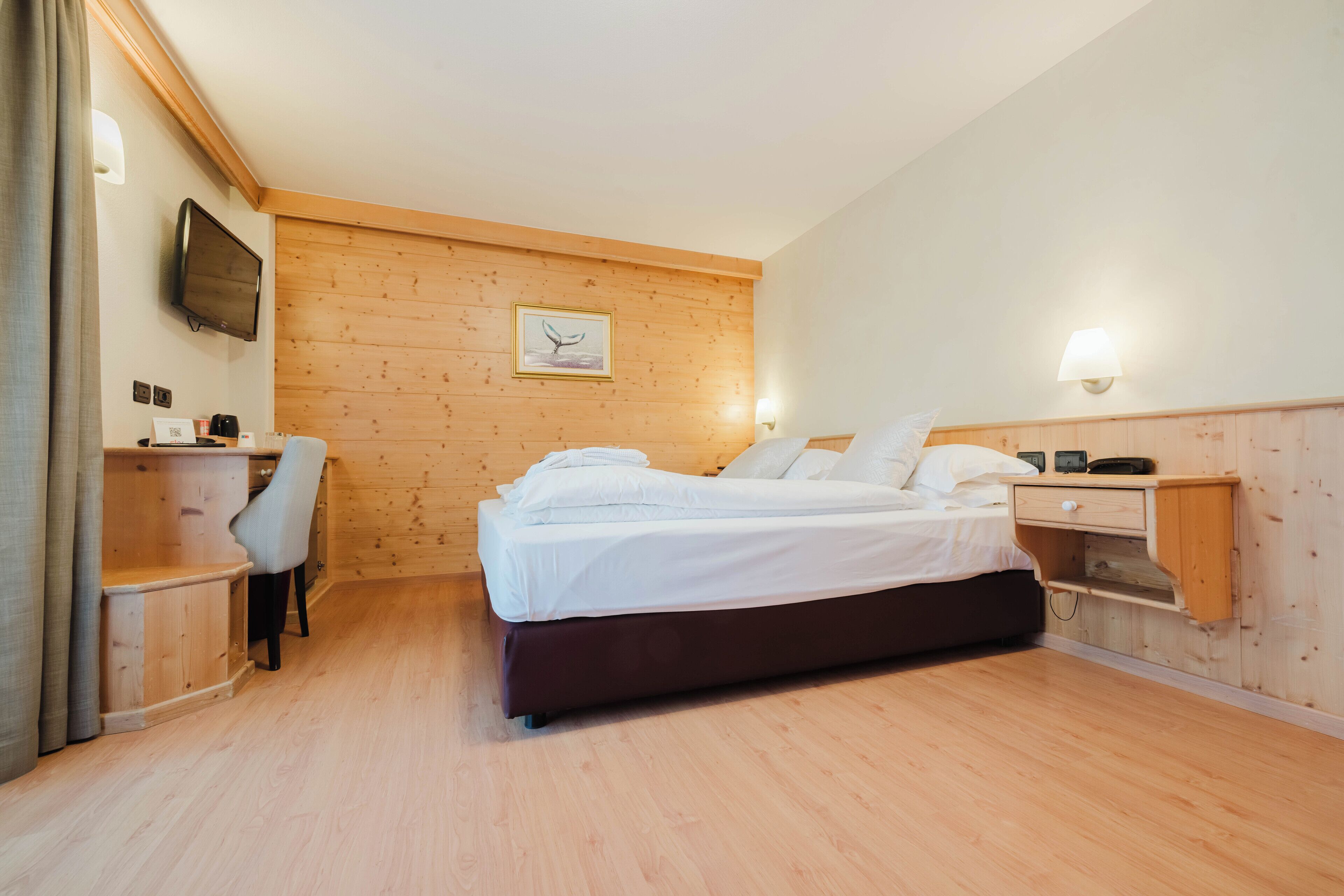 Habitación de hotel con suelo de madera, cama con ropa blanca, escritorio con silla, televisor montado en la pared y un cuadro enmarcado.