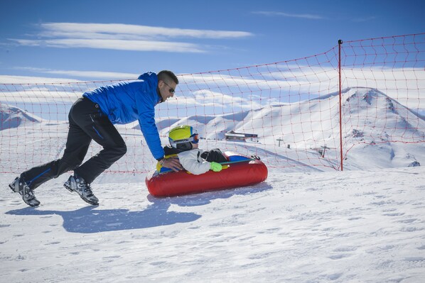 A person in a blue jacket assisting another person on a red sled on a snowy slope with a clear sky and mountains in the background.