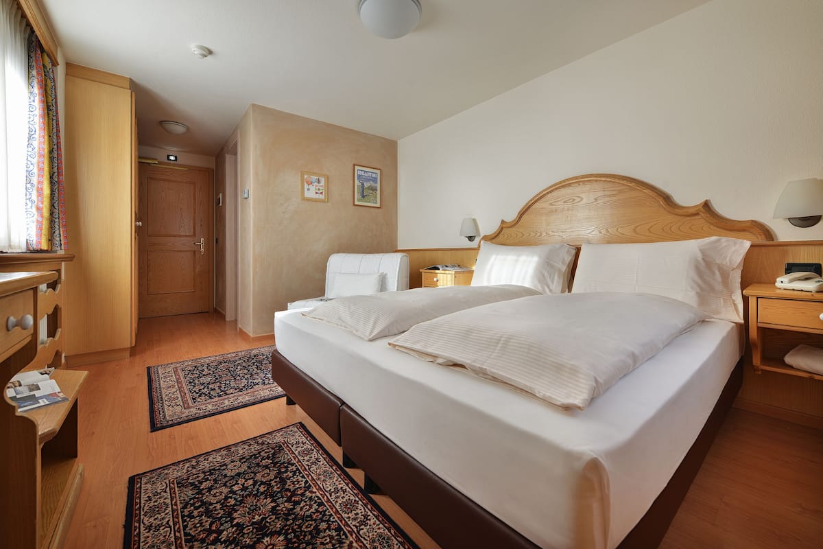 A hotel room with a large bed, wooden headboard, two bedside tables, a nightstand, a desk with a chair, a wardrobe, and a door leading to another room.