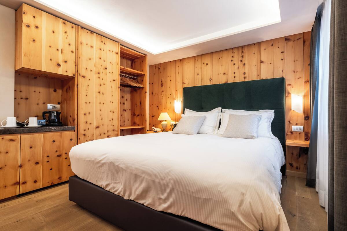 A hotel room with a large bed, wooden panel walls, a built-in wardrobe, and a small desk with a lamp.