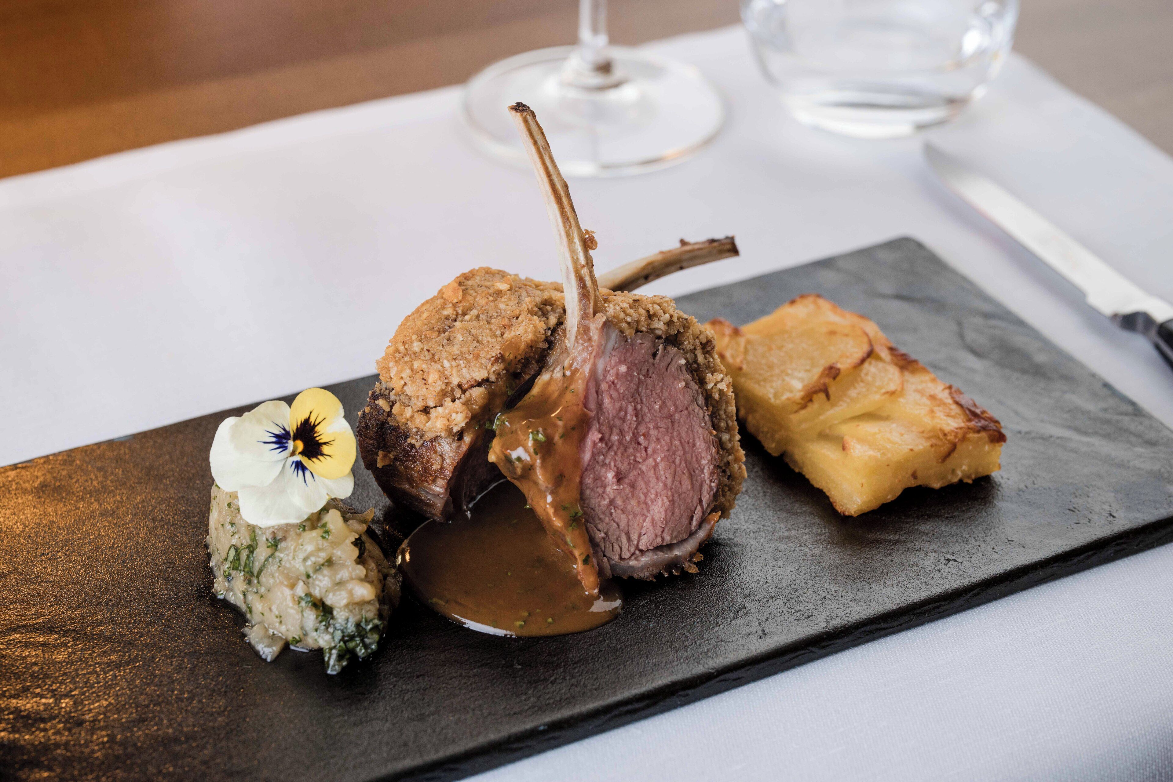 A plated dish with a lamb chop, a side of mashed potatoes, and a small flower garnish.