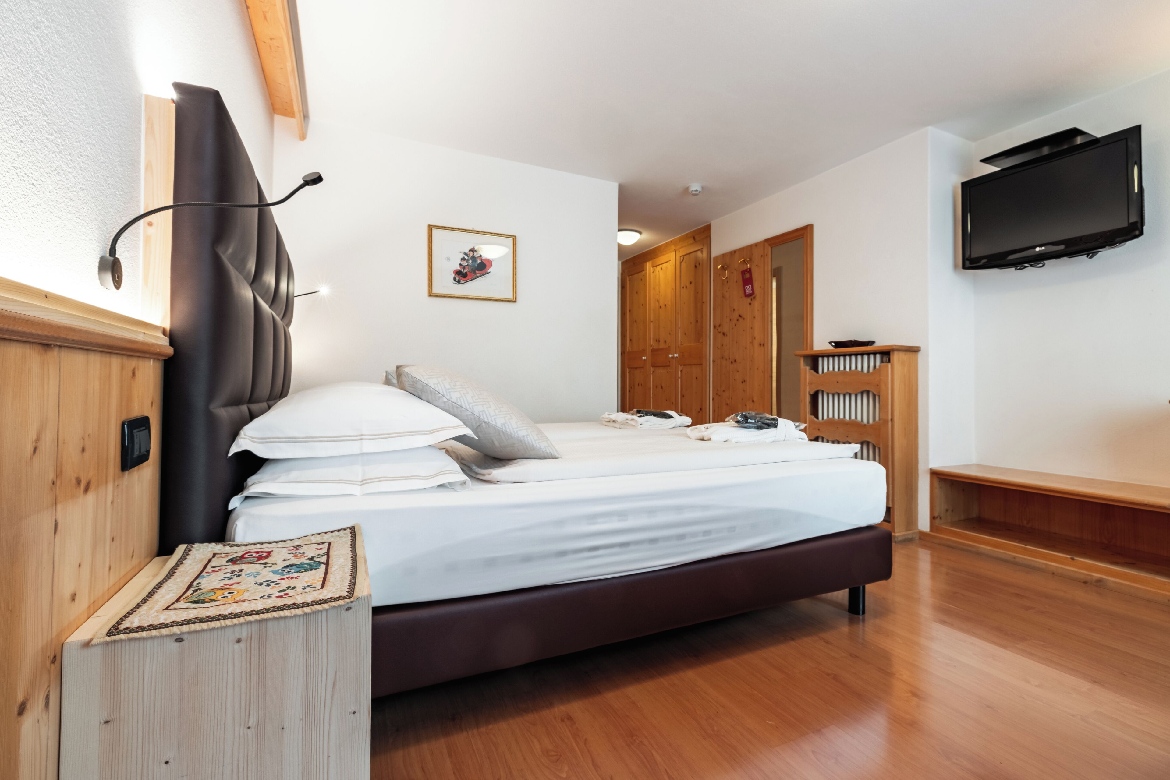 A bedroom with a bed, wooden headboard, a bedside table, a television mounted on the wall, and a wardrobe.