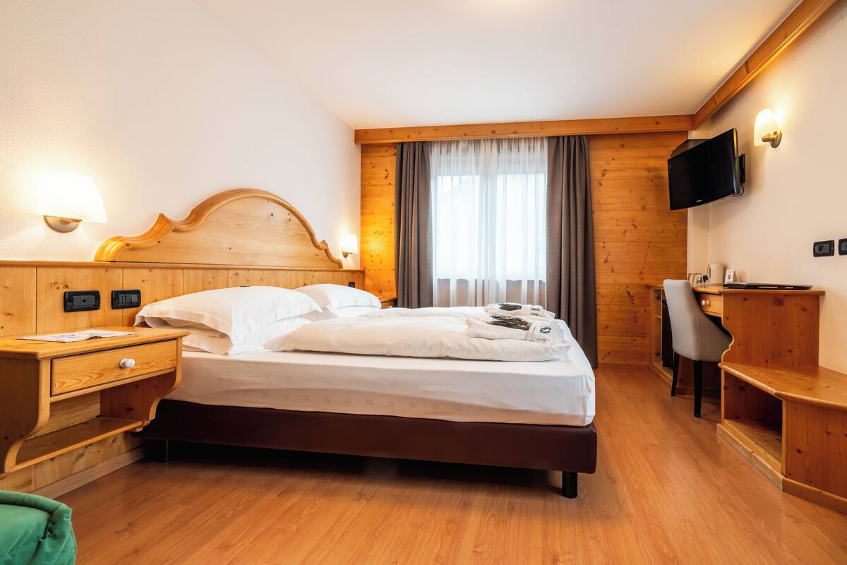 A hotel room with a wooden headboard, a bed, a desk, a chair, and a television mounted on the wall.