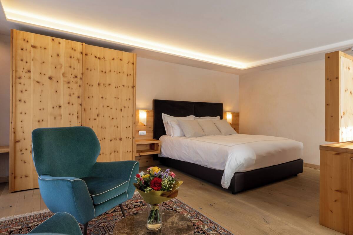 A bedroom with a bed, a blue chair, a wooden wardrobe, and a small table with a vase of flowers.