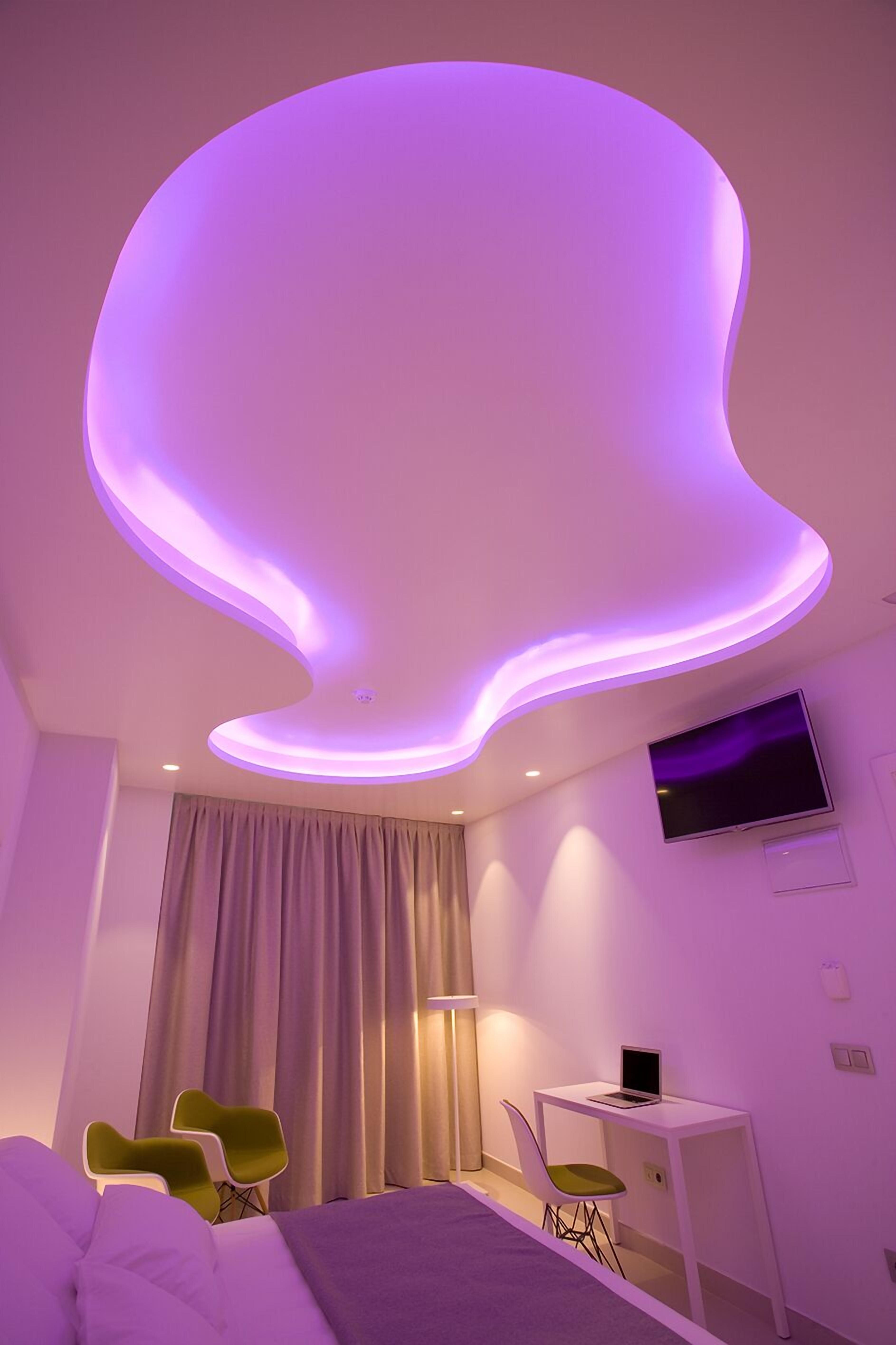 A modern bedroom with a unique ceiling design, a bed, a desk with a laptop, and a TV.