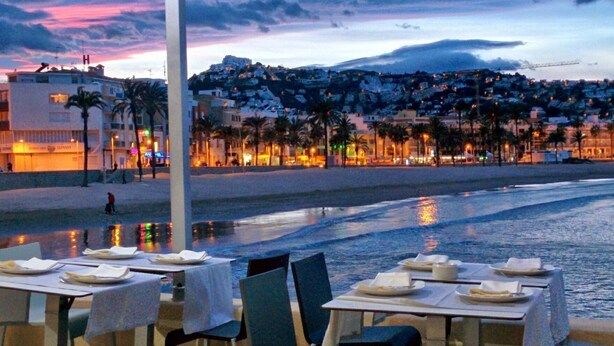 Un área de comedor al aire libre con mesas y sillas, con vistas a una playa y al horizonte urbano al anochecer.