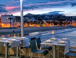 Un área de comedor al aire libre con mesas y sillas, con vistas a una playa y al horizonte urbano al anochecer.