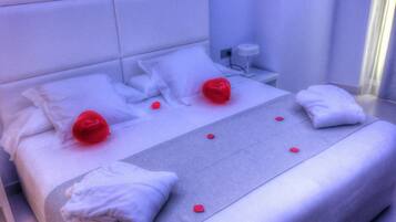 Una cama bien tendida con sábanas blancas y almohadas rojas, una mesita de noche con una lámpara y una pequeña mesita blanca.
