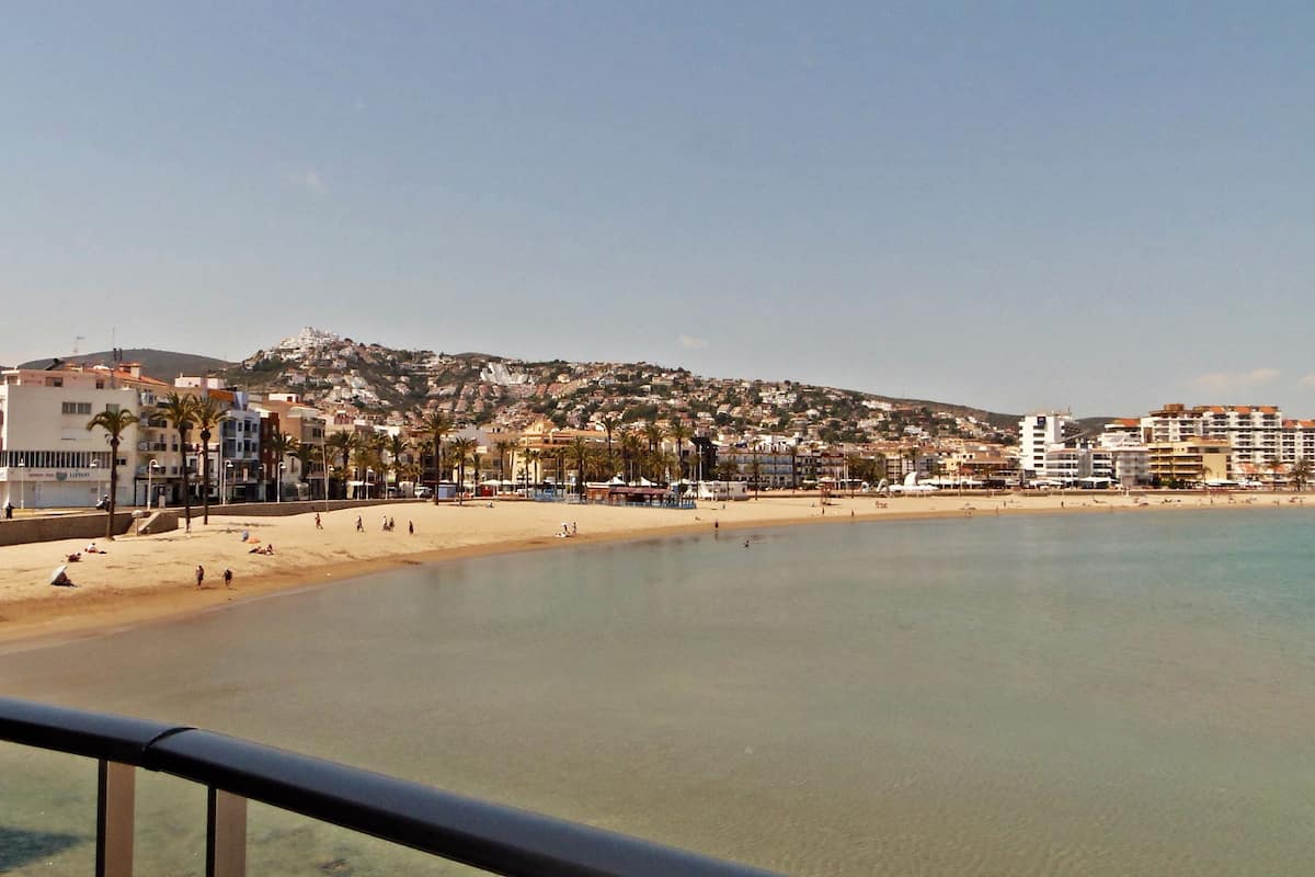 A coastal view with a clear beach, buildings in the background, and a metal railing in the foreground.