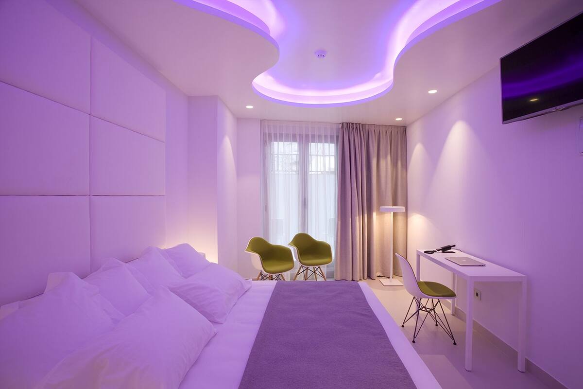A modern hotel room with a large bed, two green chairs, a desk with a laptop, and a TV mounted on the wall.