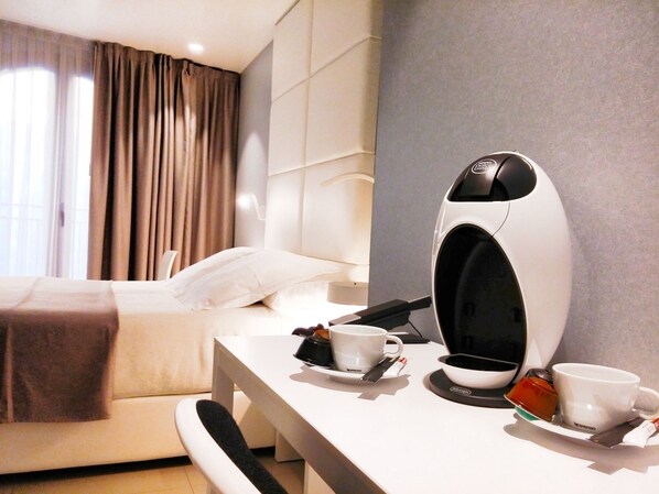 Una habitación de hotel moderna con una cama bien hecha, un escritorio blanco con cafetera, y una taza de café sobre la mesa.