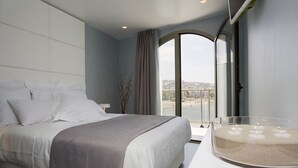 Una habitación de hotel moderna con una cama grande, una mesa de centro de vidrio con velas y vistas a un paisaje urbano a través de un ventanal amplio.