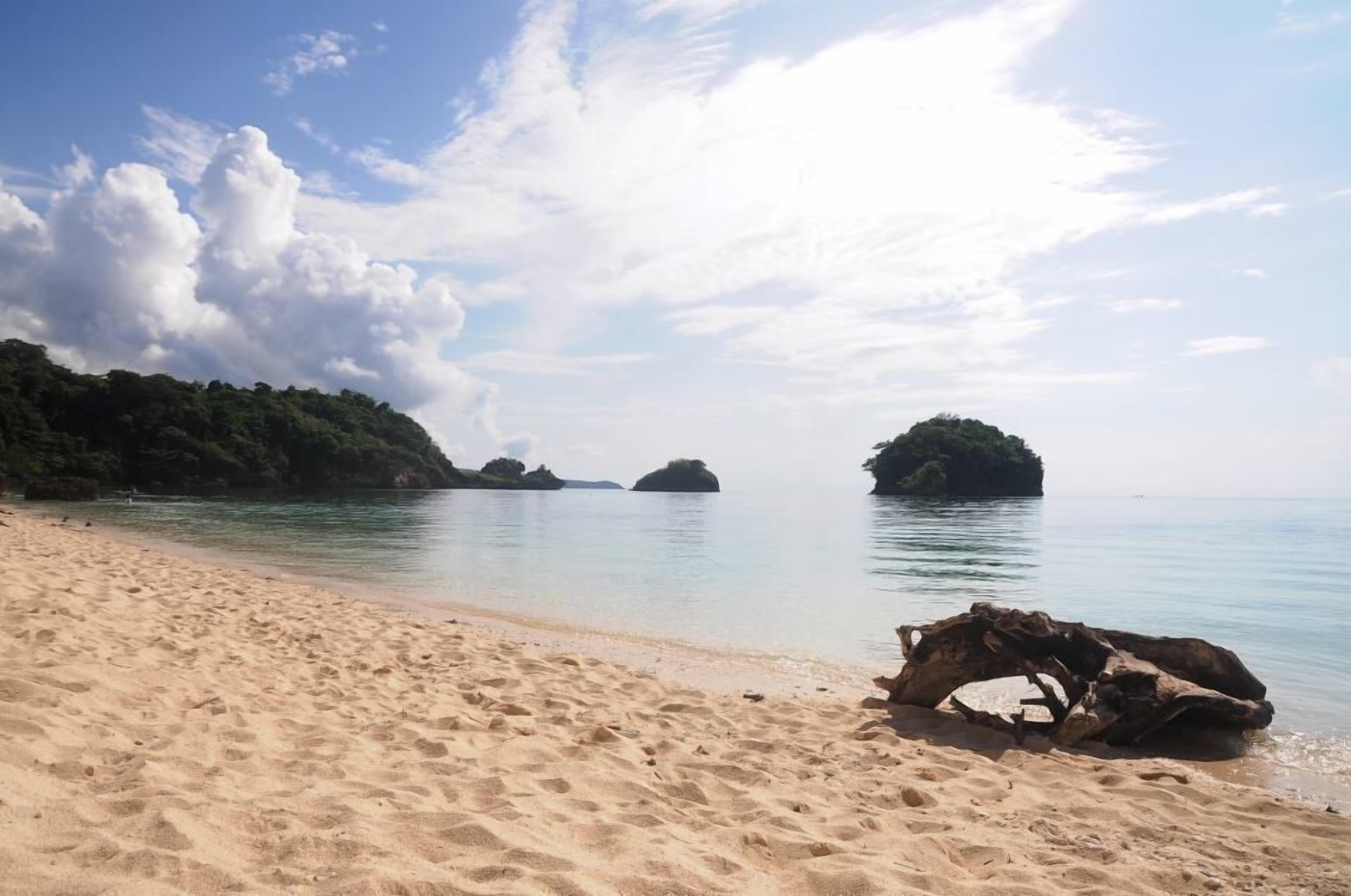 A sandy beach with clear water and a large rock formation in the distance.