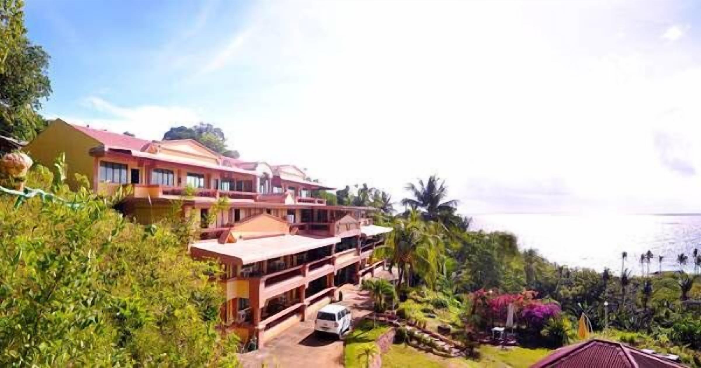A multi-story building with a red roof, surrounded by lush greenery and a view of the ocean.