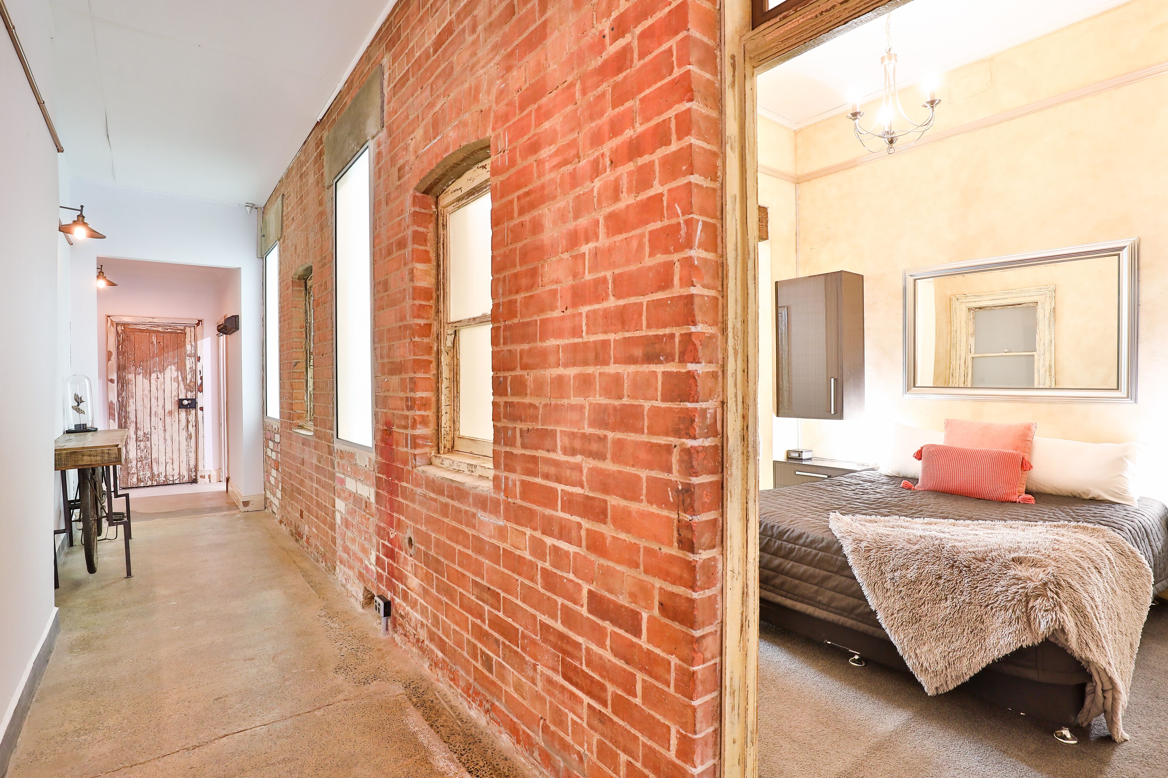 A narrow hallway with exposed brick walls, a wooden table, and a bed with a brown blanket and a red pillow.