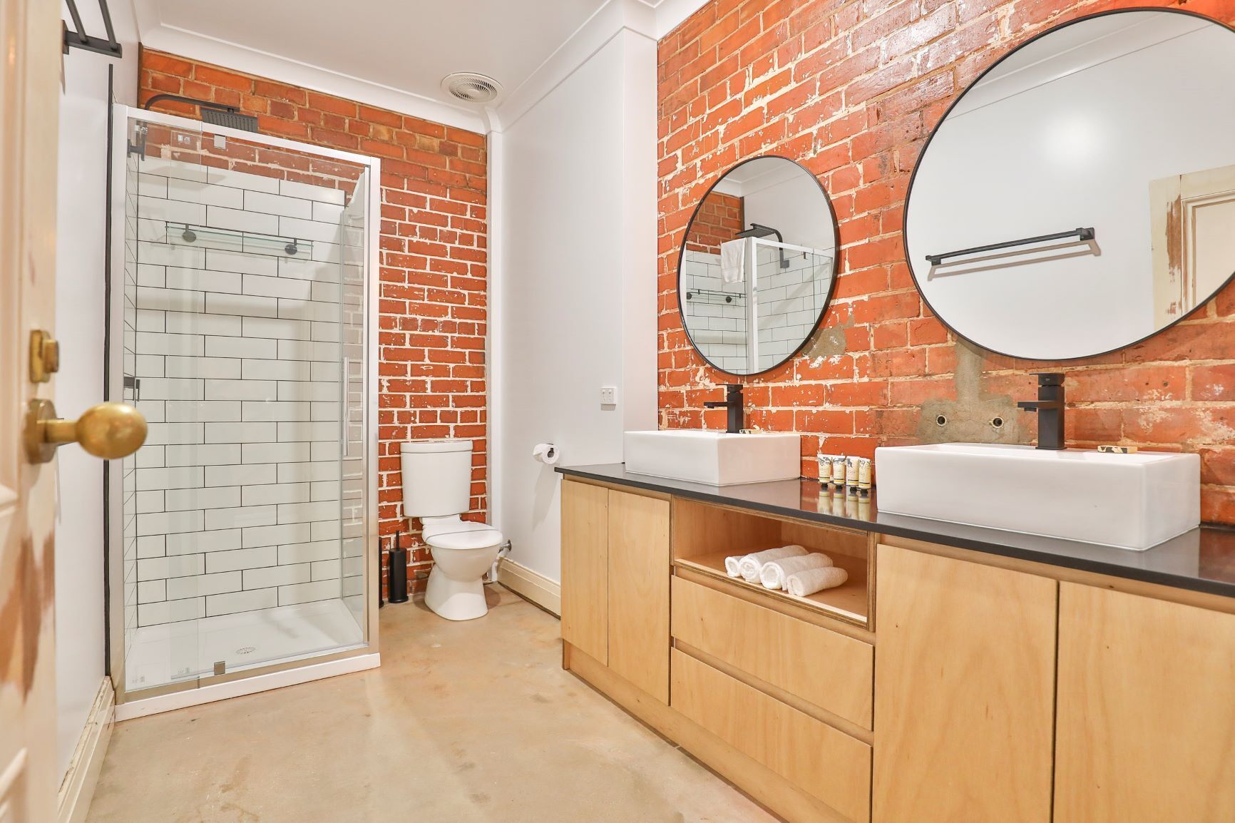 Un baño con pared de ladrillo, cabina de ducha de vidrio, inodoro y muebles de madera con encimera negra.