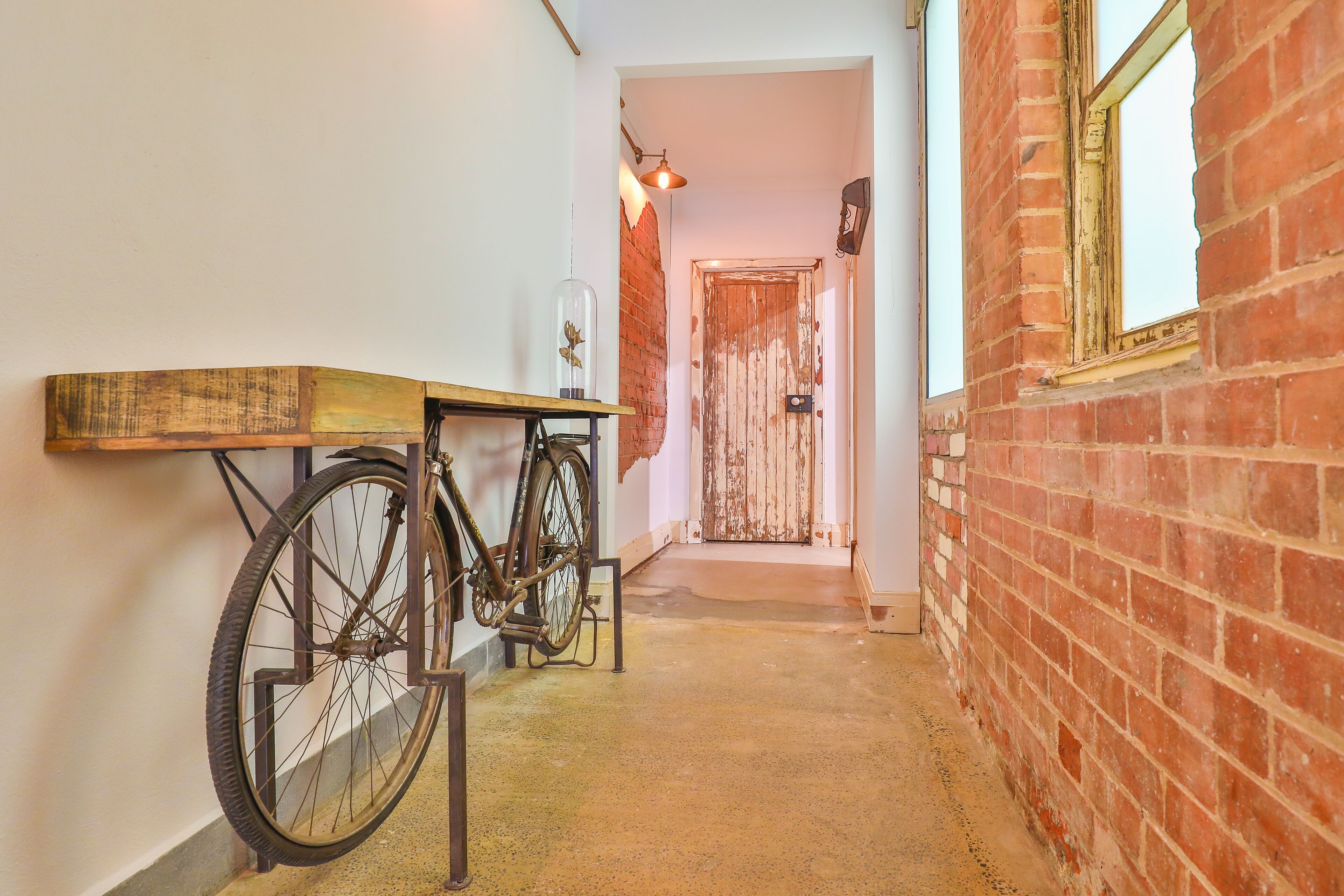 A narrow hallway with a bicycle leaning against a wall, a wooden shelf, and a rustic brick wall.