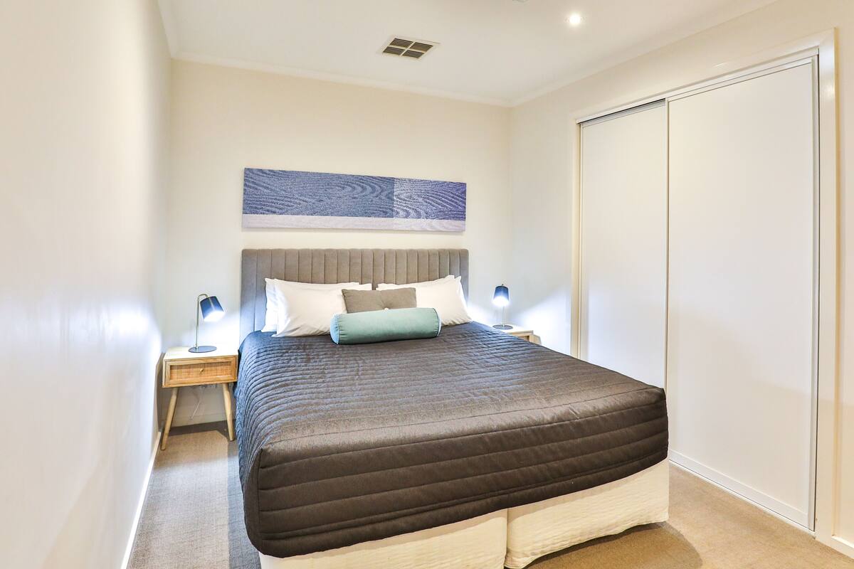 A neatly arranged bedroom with a large bed, a bedside table with a lamp, a wall-mounted artwork, and a wardrobe.