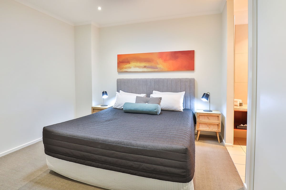 A modern bedroom with a large bed, a headboard, two bedside tables with lamps, and a wall-mounted abstract painting.