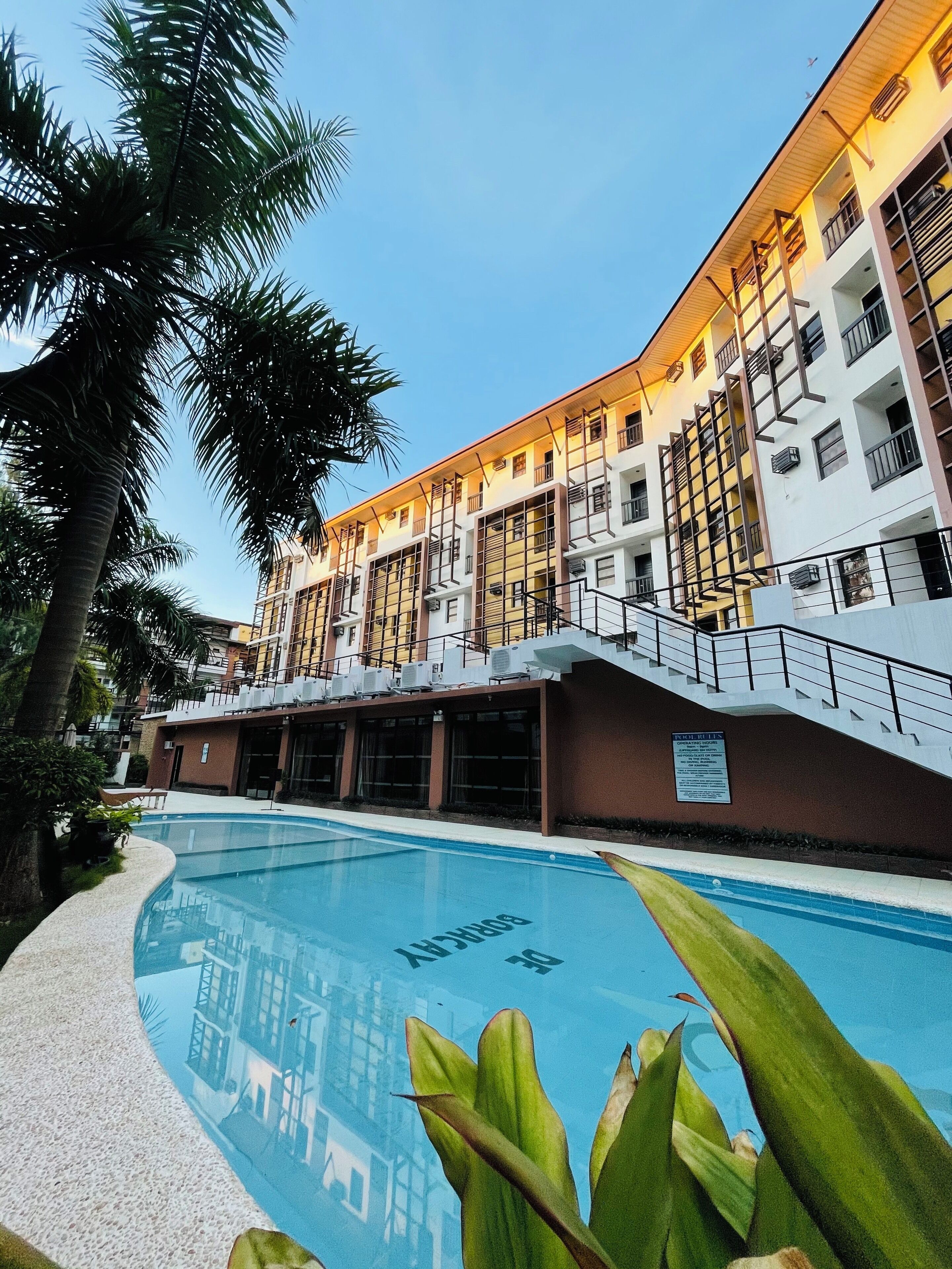 Un hôtel à plusieurs étages doté d’une piscine, de palmiers et d’un panneau indiquant les règles à respecter au bord de la piscine.