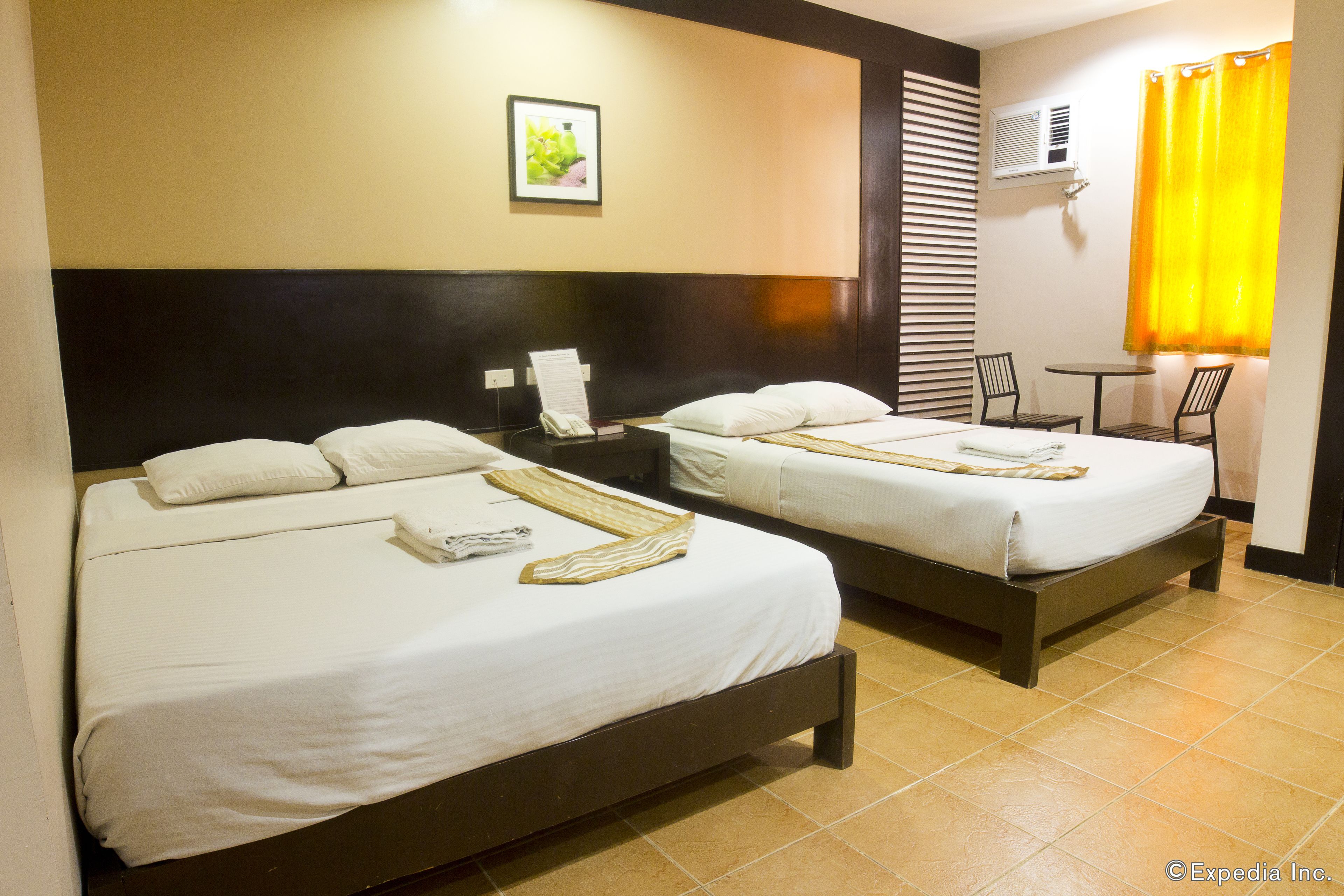 A hotel room with two beds, a small table, chairs, a wall-mounted air conditioner, and a window with yellow curtains.