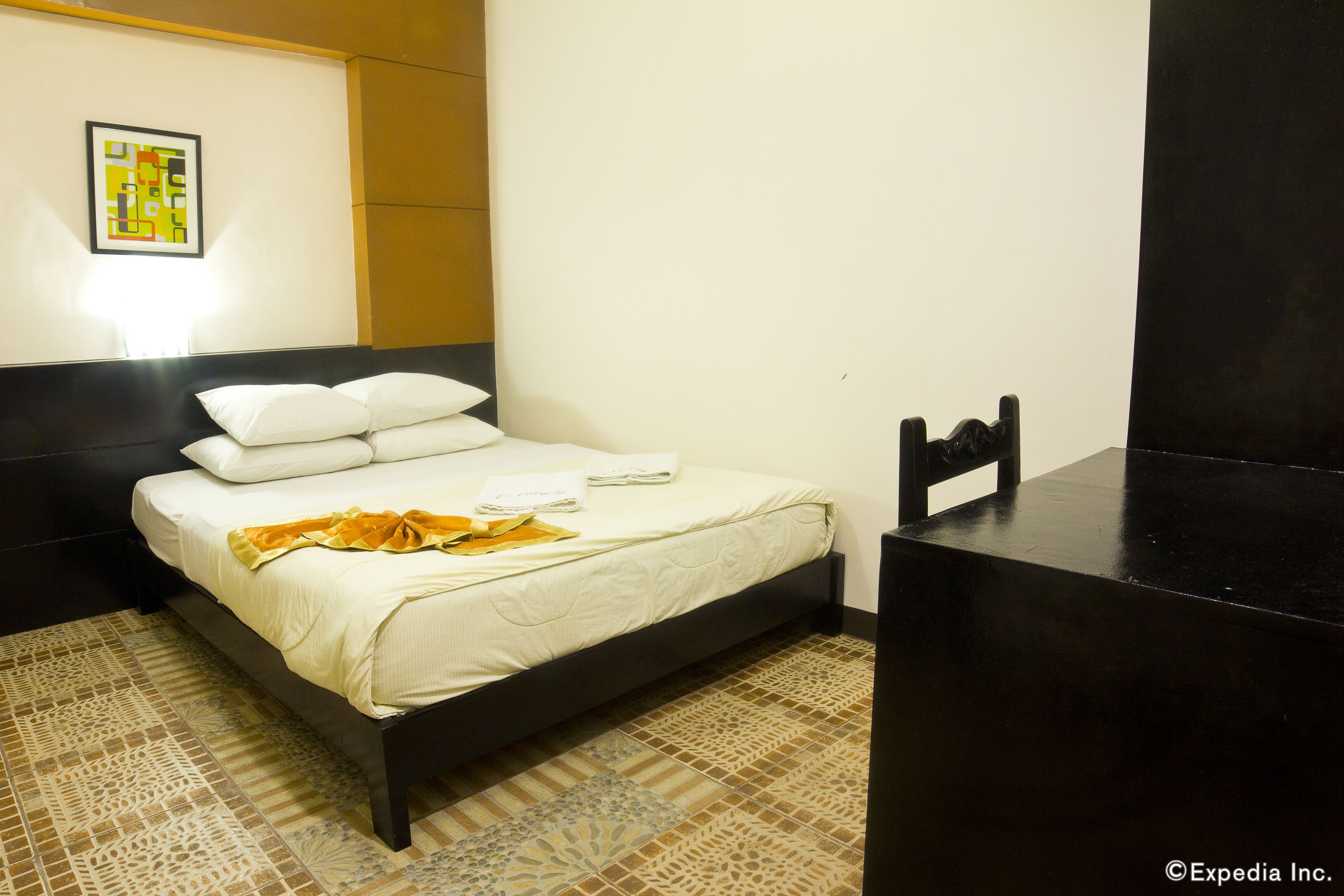 A neatly made bed with white bedding and a yellow blanket, a black headboard, and a patterned rug on the floor.