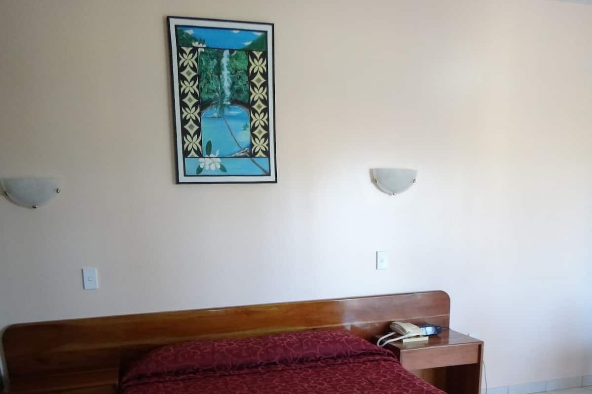 A hotel room with a wooden bed, red patterned bedspread, two towels on the bed, a nightstand with a phone, and a framed picture on the wall.