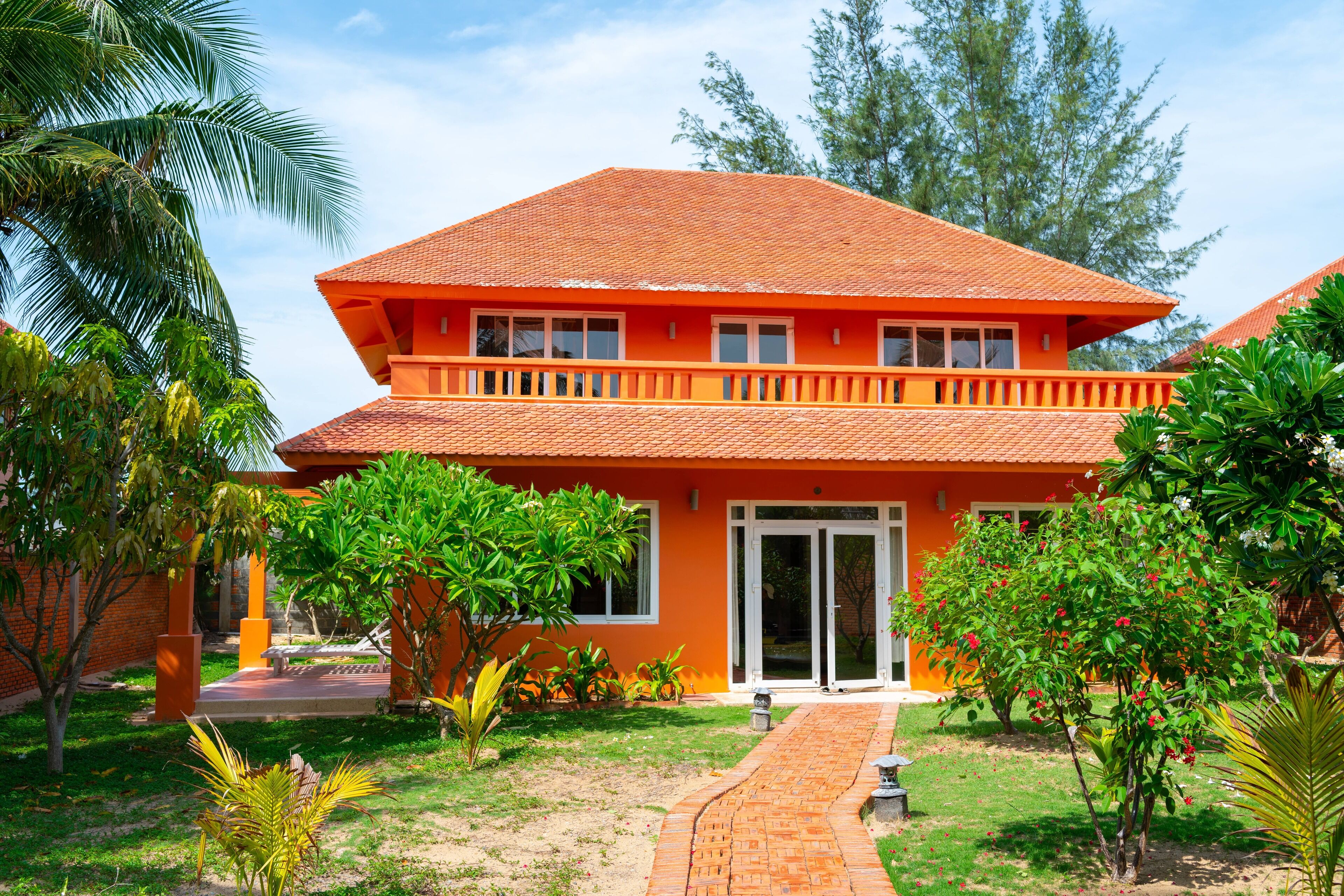 A two-story building with a red roof and white-framed windows, surrounded by greenery and a paved pathway.