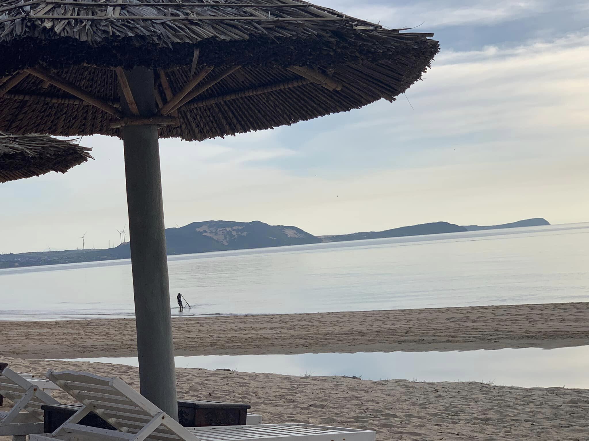 A beach with a thatched umbrella, lounge chairs, and a calm sea with distant mountains.
