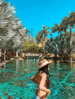 Una mujer en traje de baño y sombrero de paja, de pie en una piscina cristalina rodeada de palmeras y vegetación tropical.