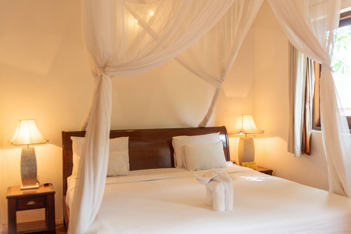 A bed with white linens, a wooden headboard, and a canopy. There are two bedside lamps and a nightstand with a lamp.