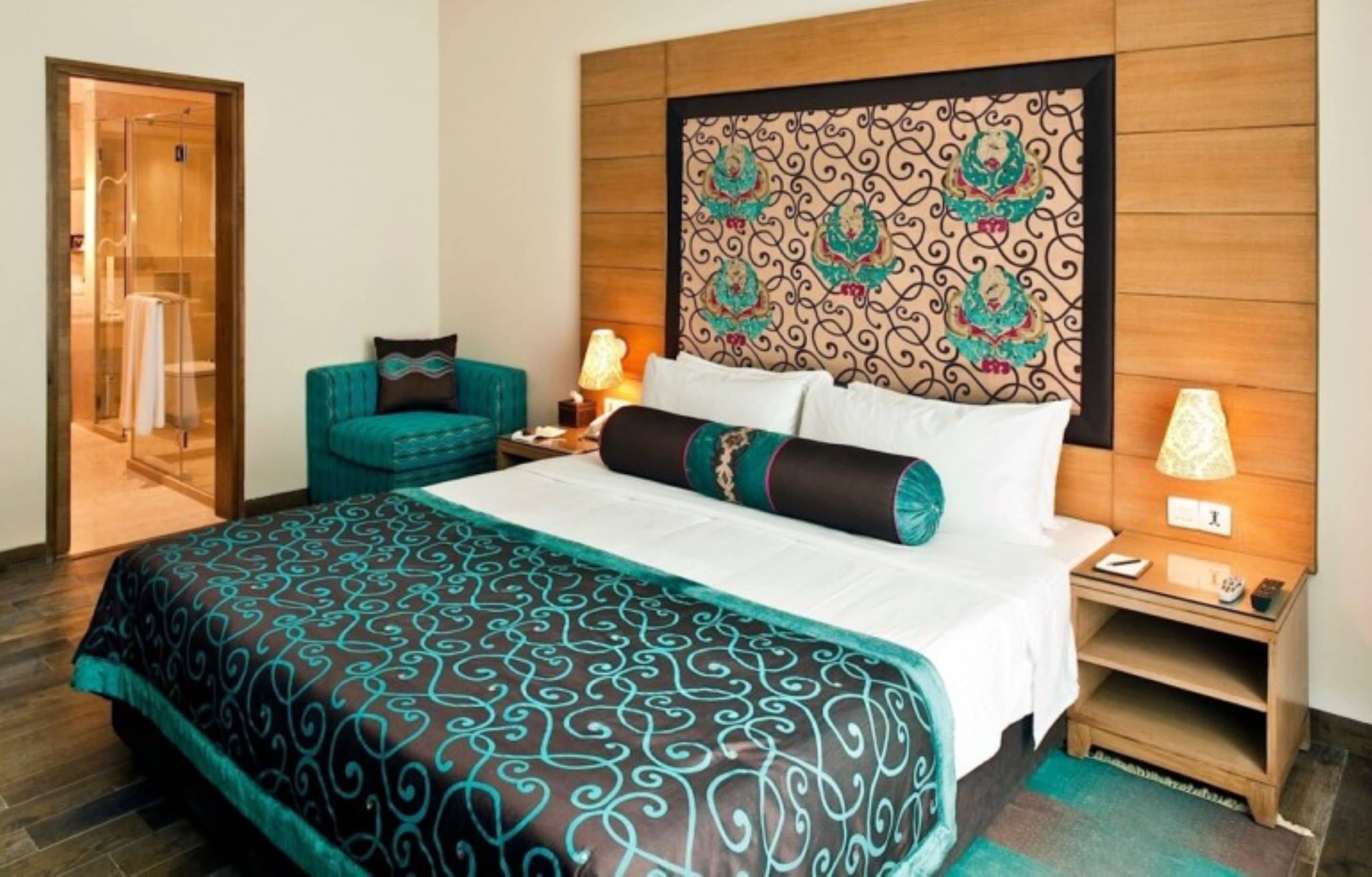 Habitación de hotel con una cama grande, cabecera de madera, un sillón azul turquesa, lámparas de noche y un box de ducha de vidrio.