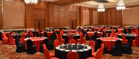 A large banquet hall with round tables, red and black chairs, chandeliers, and a red carpeted floor.